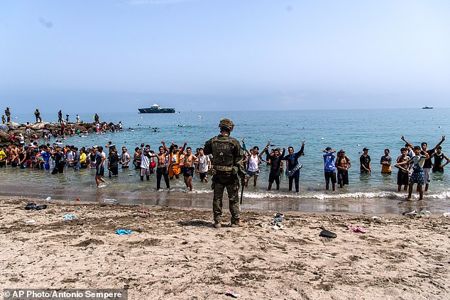 A group of migrants pictured standing in the water and waving as a Spanish army soldier keeps guard